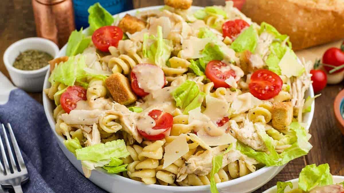 A bowl of pasta salad with rotini, lettuce, cherry tomatoes, croutons, shredded chicken, and parmesan cheese, topped with creamy dressing.