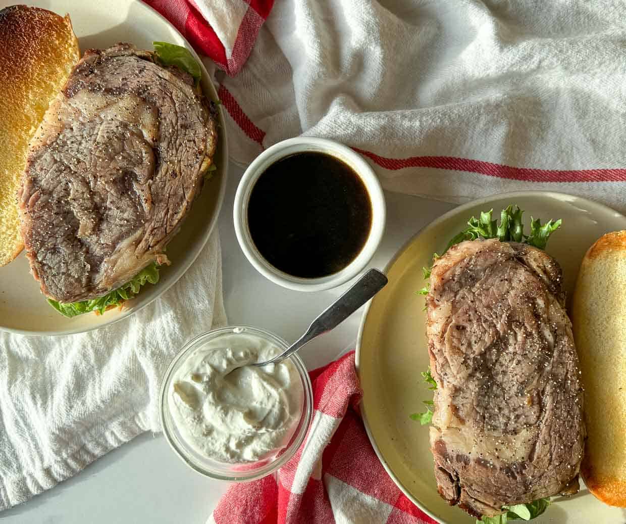 Two plates with sandwiches containing thick slices of roast beef and lettuce, a bowl of creamy sauce, and a cup of dark dipping sauce on a white surface with red-striped towels.