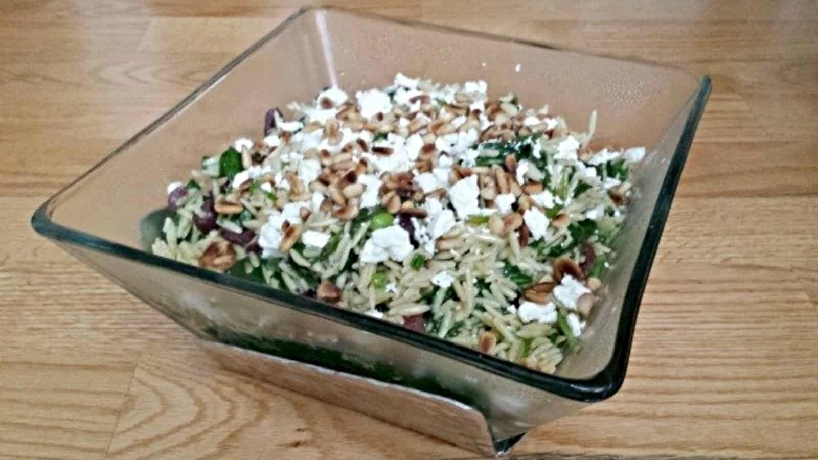 A square glass bowl filled with orzo salad, topped with crumbled feta cheese, pine nuts, herbs, and chopped vegetables, placed on a wooden surface.