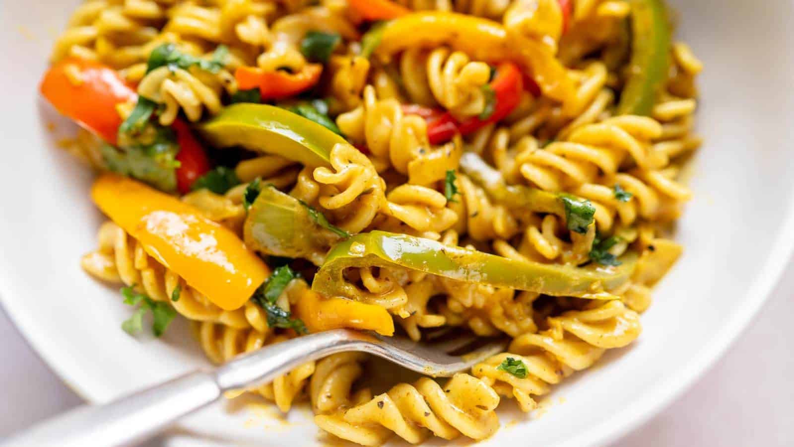 A close-up of rotini pasta mixed with sliced red and yellow bell peppers, green herbs, and a fork on a white plate.