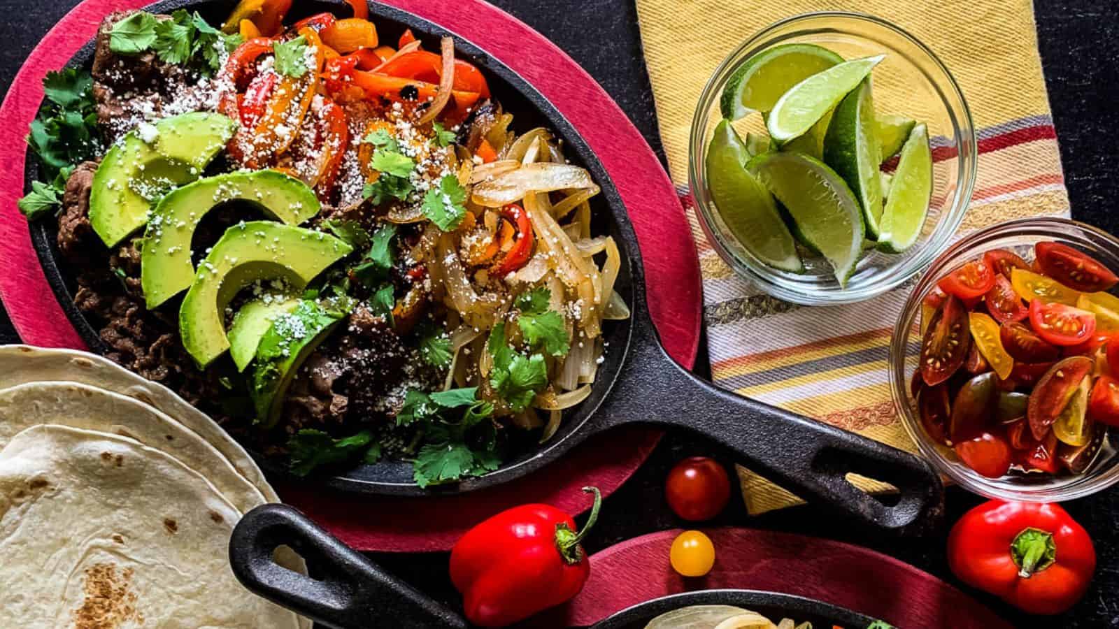 A skillet with sliced avocado, sautรฉed peppers, onions, ground beef, and cilantro, served with tortillas, lime wedges, and cherry tomatoes on the side.