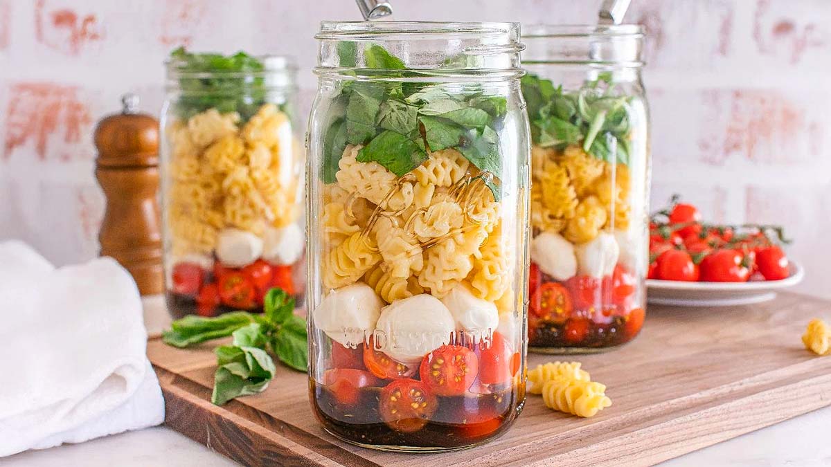 Three glass jars are filled with cherry tomatoes, mozzarella balls, pasta, spinach, and basil, arranged in layers, sitting on a wooden board with utensils and fresh ingredients nearby.