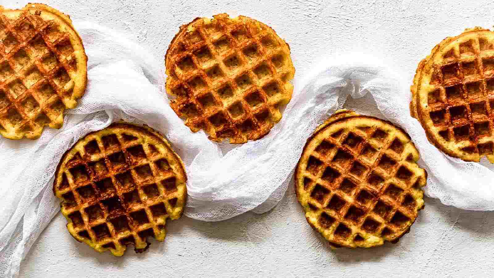 Five golden brown waffles are arranged in a row on a light surface, partially resting on a white cloth.