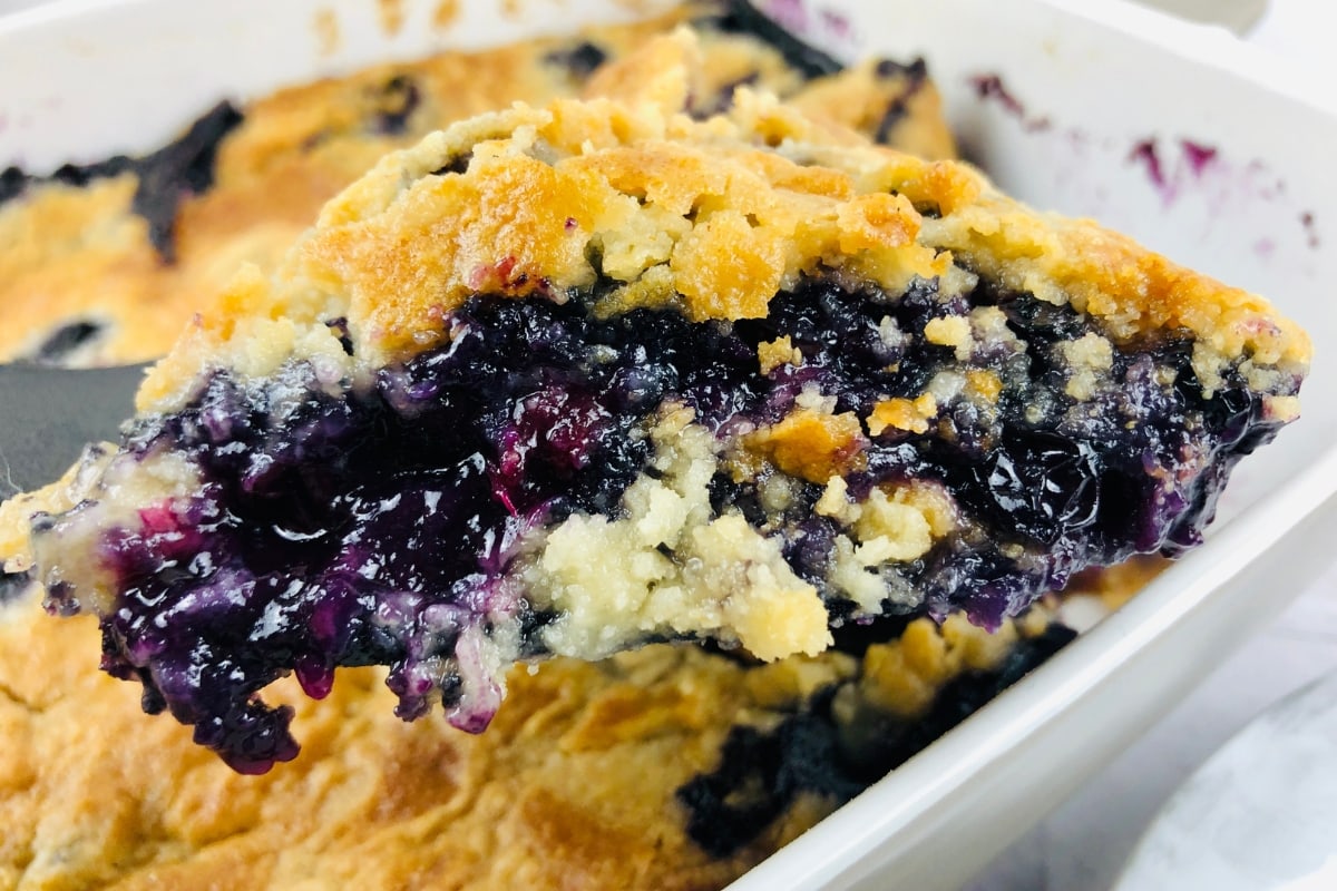 A close-up of a serving of blueberry cobbler with a golden-brown crust and juicy blueberry filling on a spatula, above the baking dish.