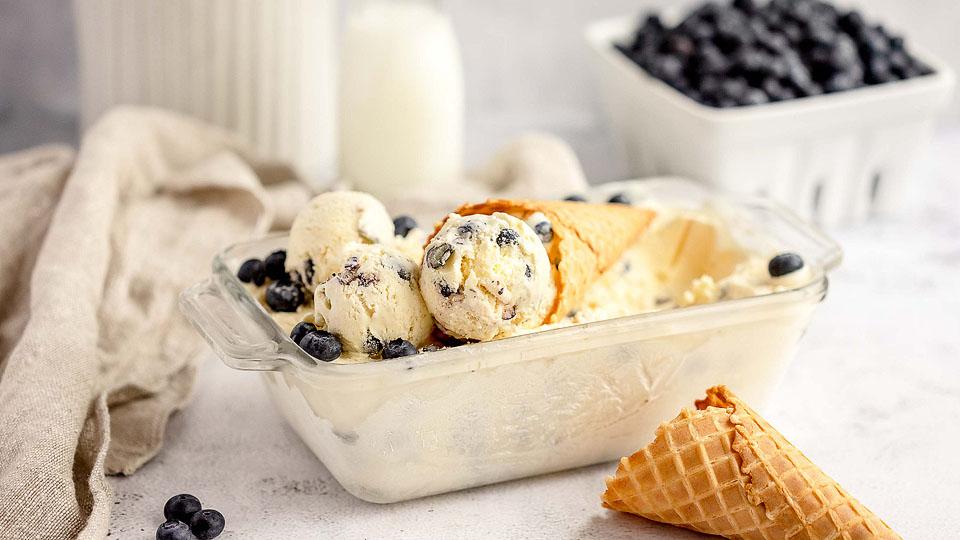 A glass dish with scoops of vanilla ice cream mixed with blueberries, an ice cream cone, and loose blueberries on a white surface. A broken cone and a container of blueberries are in the background.