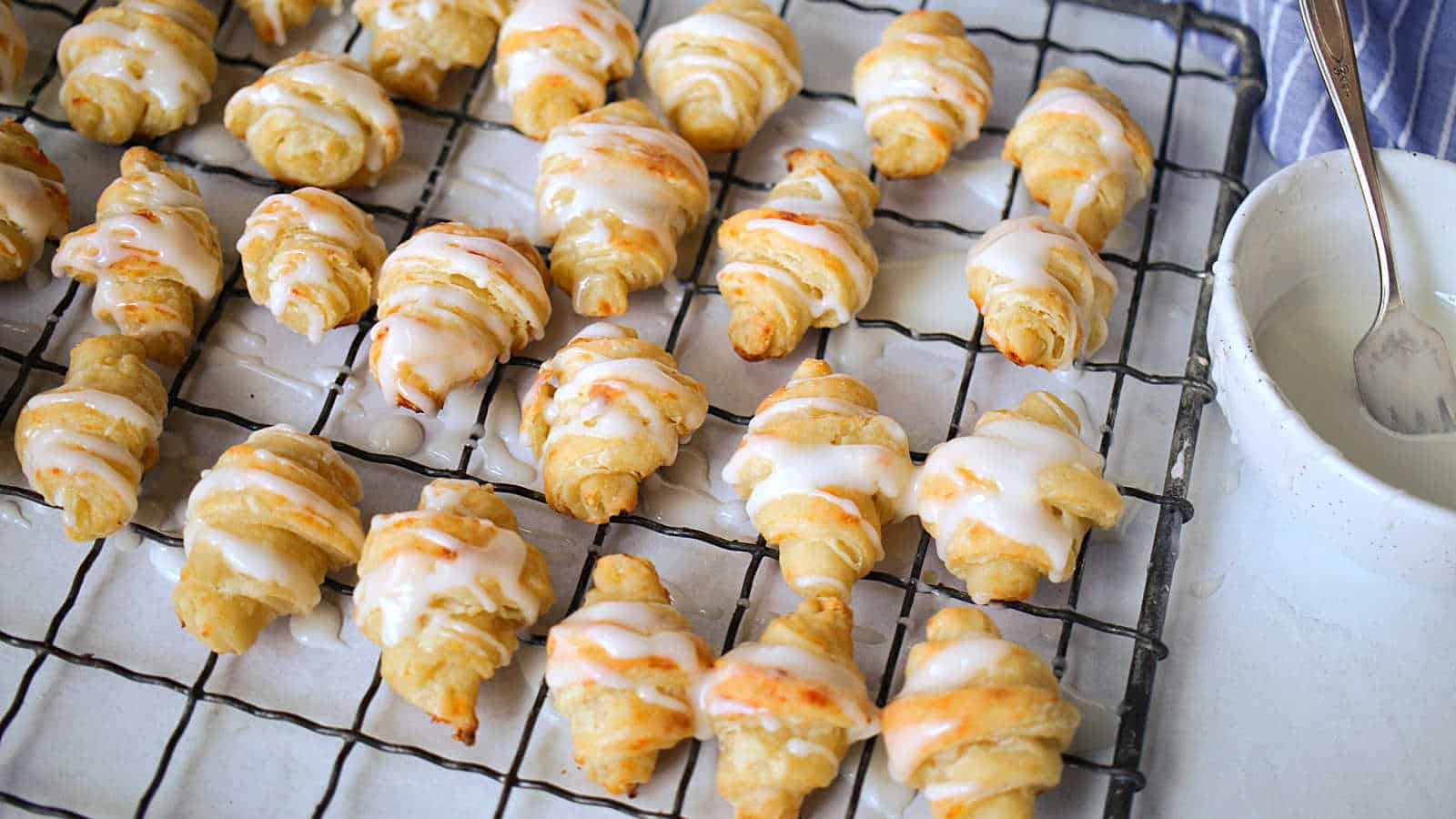 Small crescent-shaped pastries with white icing are arranged on a wire cooling rack, with a bowl and fork nearby.