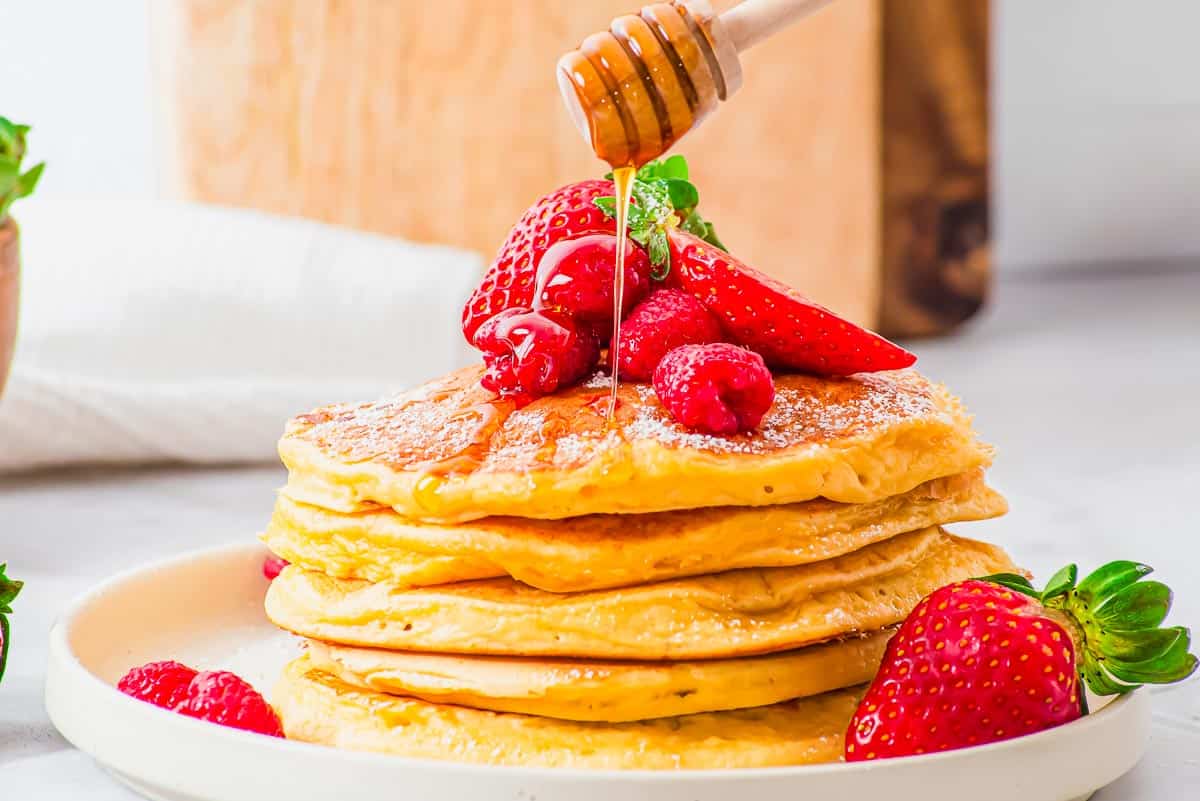 A stack of pancakes topped with strawberries and raspberries is drizzled with honey on a white plate.