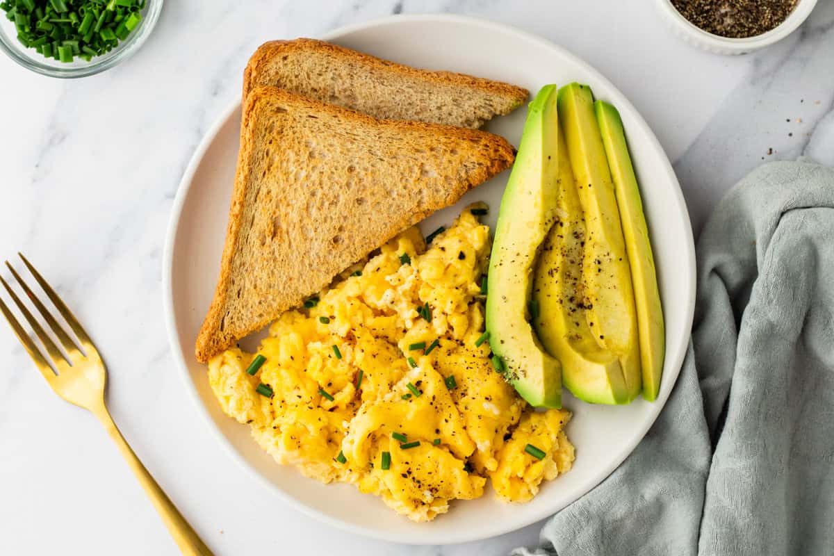 A plate with scrambled eggs topped with chives, two slices of whole wheat toast, and sliced avocado, next to a gold fork and a light gray napkin.