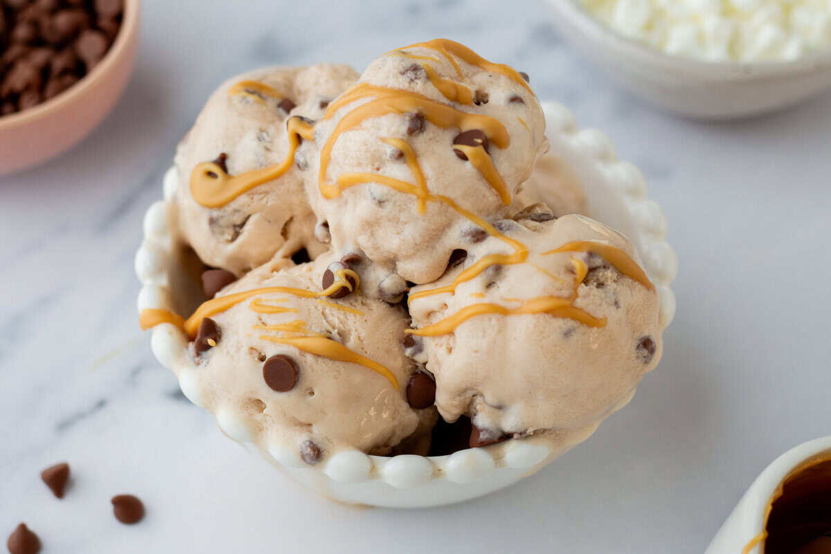 Four scoops of ice cream with chocolate chips and caramel drizzle in a white bowl, placed on a white surface with bowls of toppings in the background.