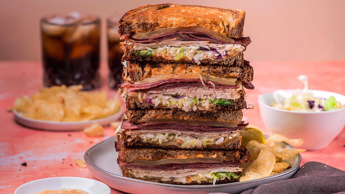 A stack of thick, deli-style sandwiches filled with layers of meat, cheese, and vegetables on rye bread, with potato chips and a drink in the background.