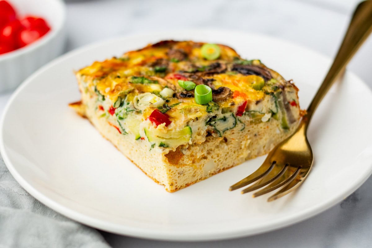 A square slice of vegetable egg casserole with visible bell peppers and green onions on a white plate, next to a gold fork.