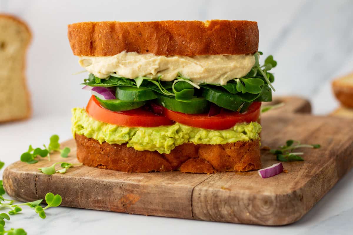 A vegetable sandwich with hummus, avocado, tomato, cucumber, spinach, red onion, and sprouts on sliced bread, placed on a wooden cutting board.