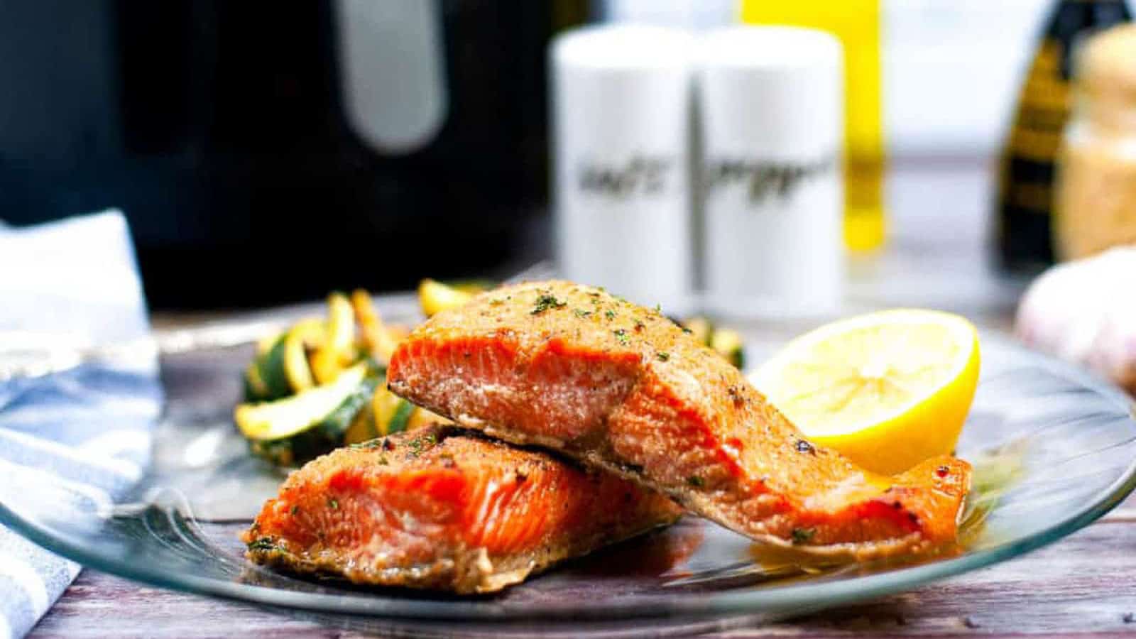 Two cooked salmon fillets stacked on a clear plate, served with grilled vegetables and a lemon wedge, with seasoning containers in the background.