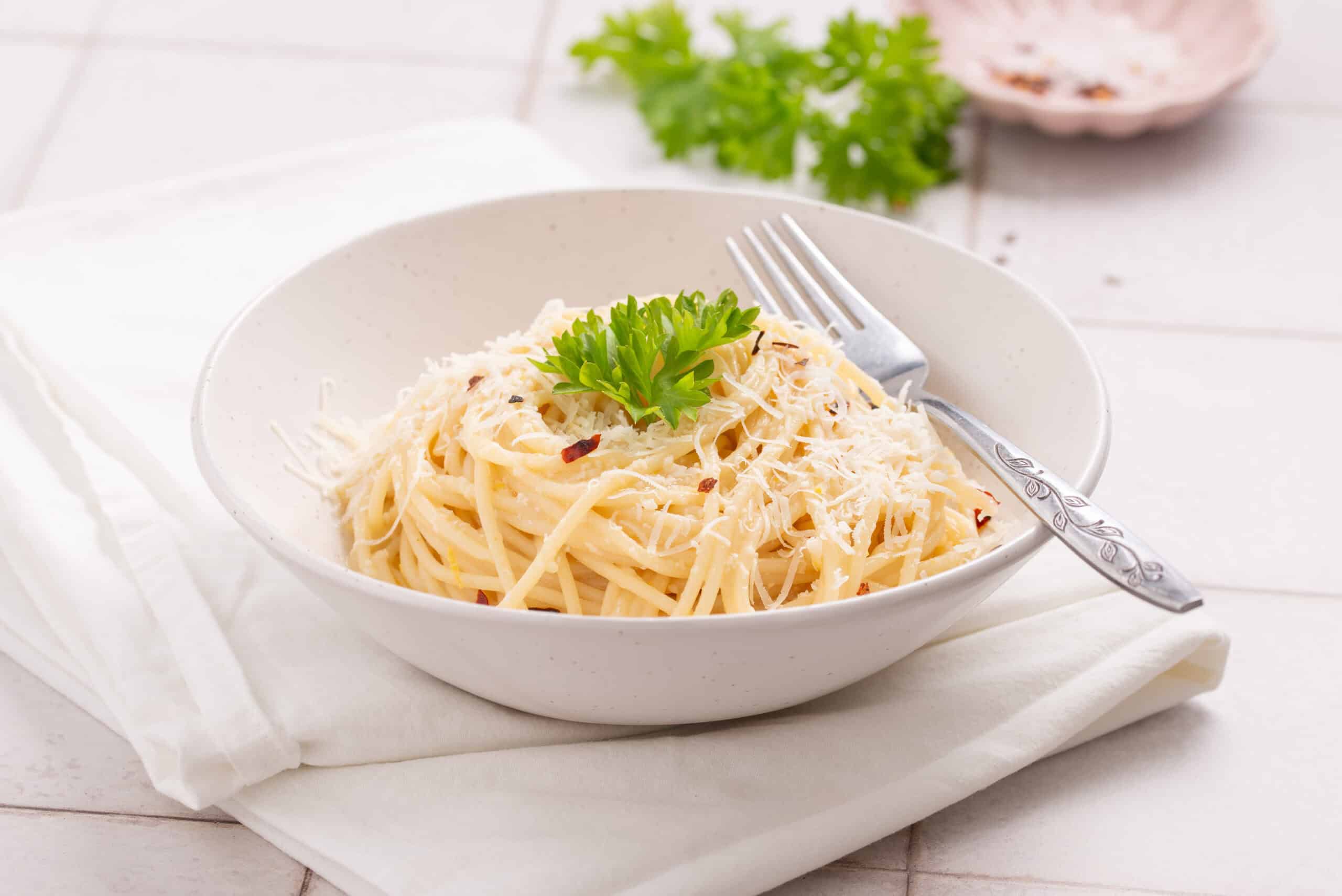 A white bowl of pasta topped with grated cheese and parsley, with a fork resting on the side, placed on a white napkin and tiled surface.