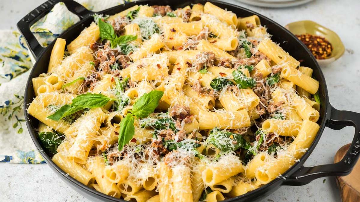 A black skillet filled with rigatoni pasta, shredded meat, spinach, grated cheese, and fresh basil leaves, on a light surface.