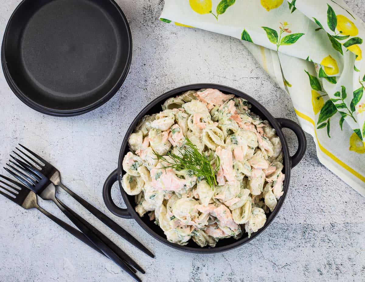 A black pot filled with creamy pasta and salmon, garnished with dill, sits on a grey surface next to black plates, cutlery, and a lemon-patterned cloth.