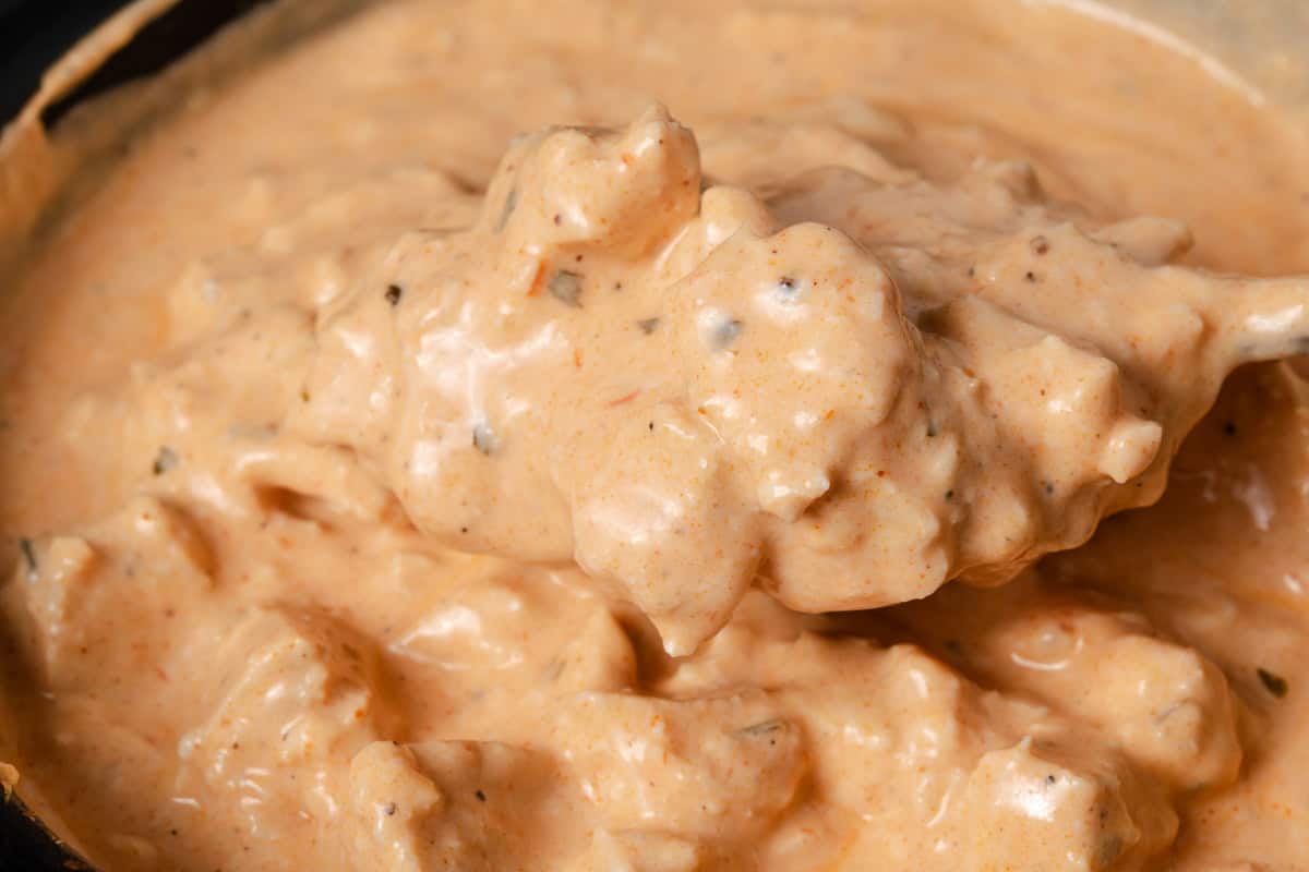 Close-up of a thick, creamy, orange-tinted dip with visible chunks and specks of seasoning, being scooped up with a spoon.