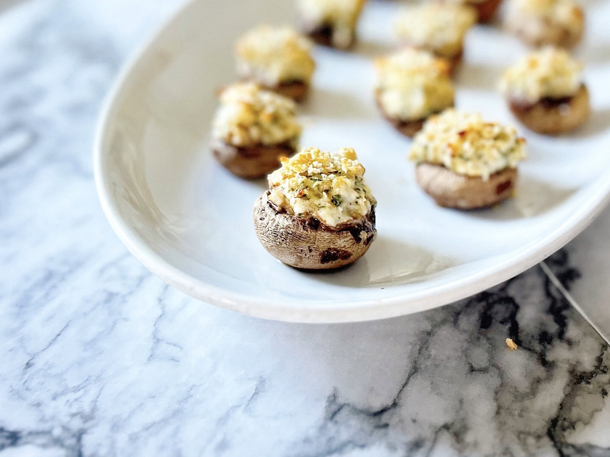 Stuffed mushrooms arranged on a white oval plate, topped with a golden breadcrumb mixture, set on a marble surface.