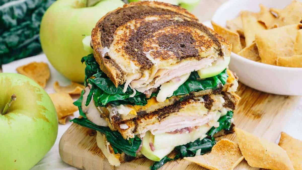 A stacked grilled sandwich with turkey, cheese, spinach, and apple slices on marbled bread, served with green apples and pita chips.