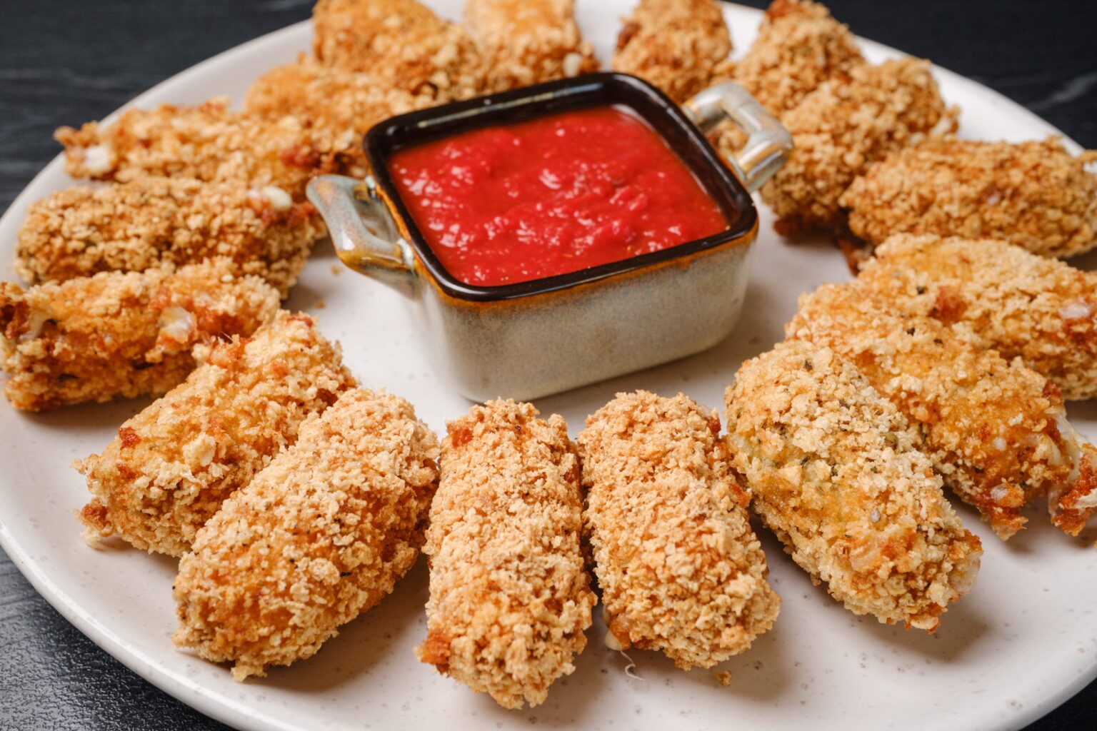 A plate of breaded, baked or fried croquettes arranged in a circle around a square dish of red dipping sauce.