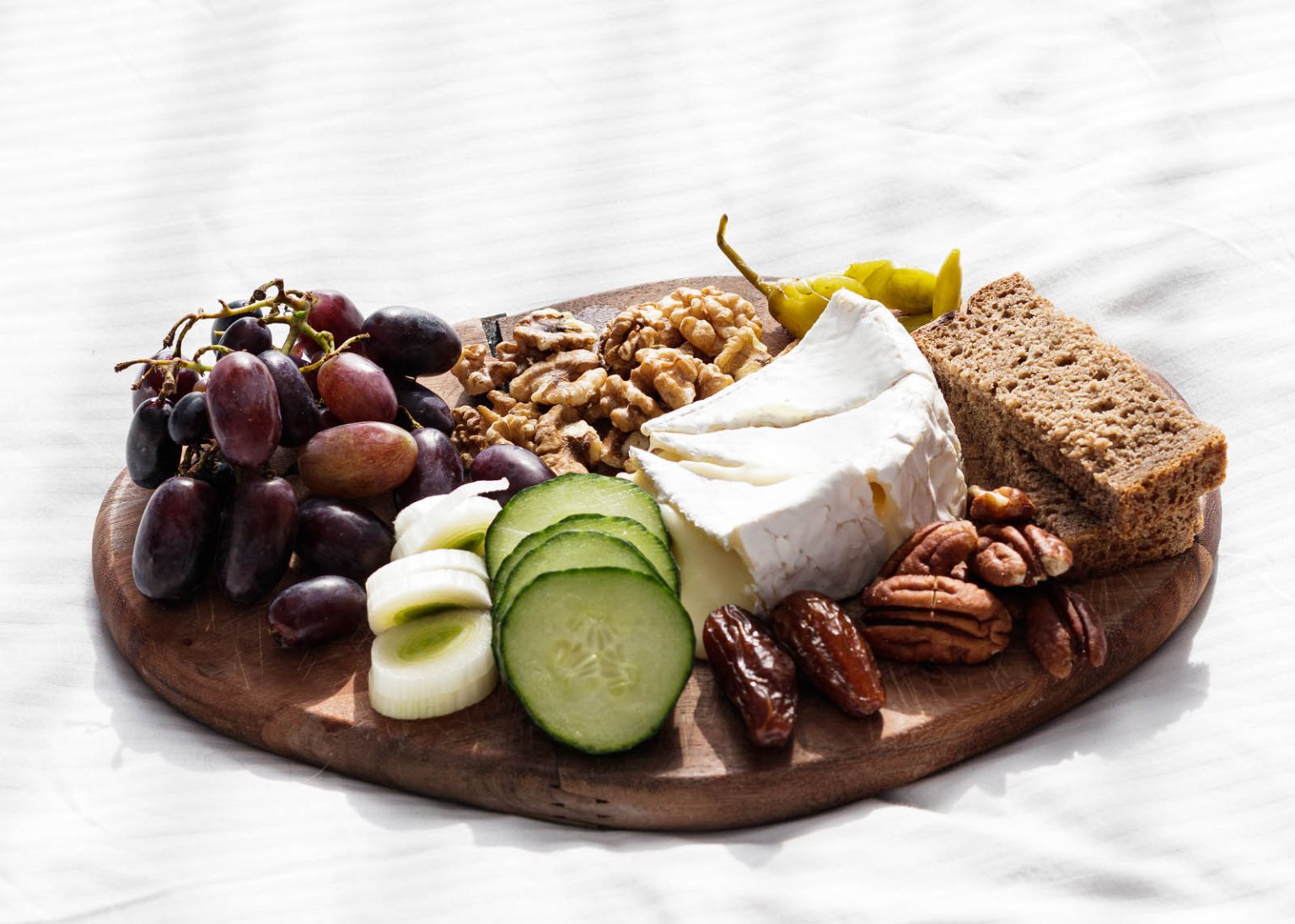 A wooden board with grapes, walnuts, Brie cheese, pecans, dates, cucumber slices, leek, brown bread, and a yellow pepper on a white surface.