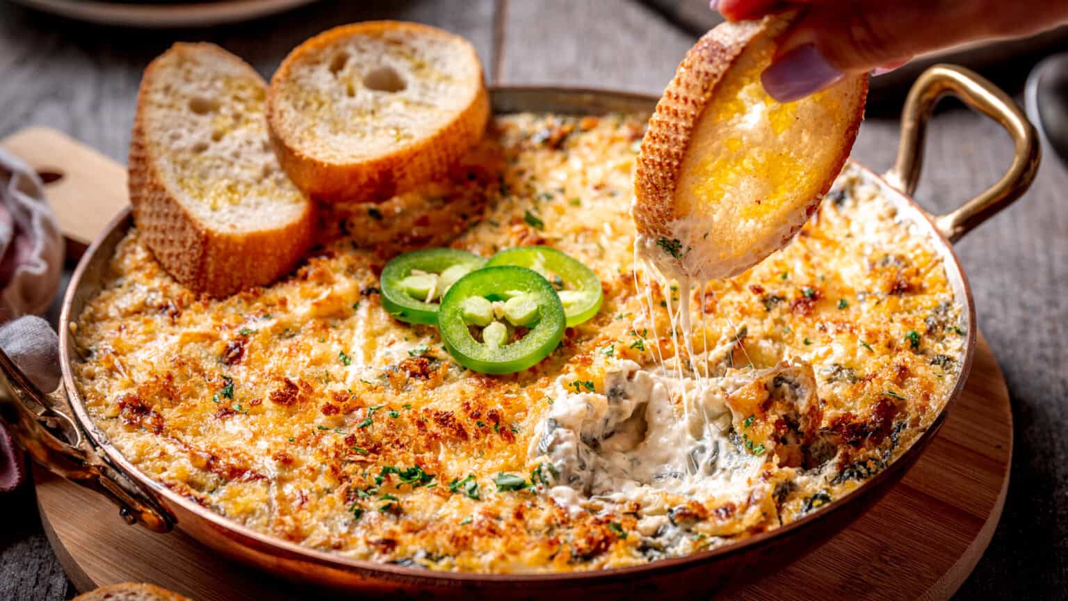 A hand dips a slice of toasted bread into a baked creamy spinach and cheese dip, garnished with jalapeรฑo slices. Other bread slices are arranged on the side of the pan.