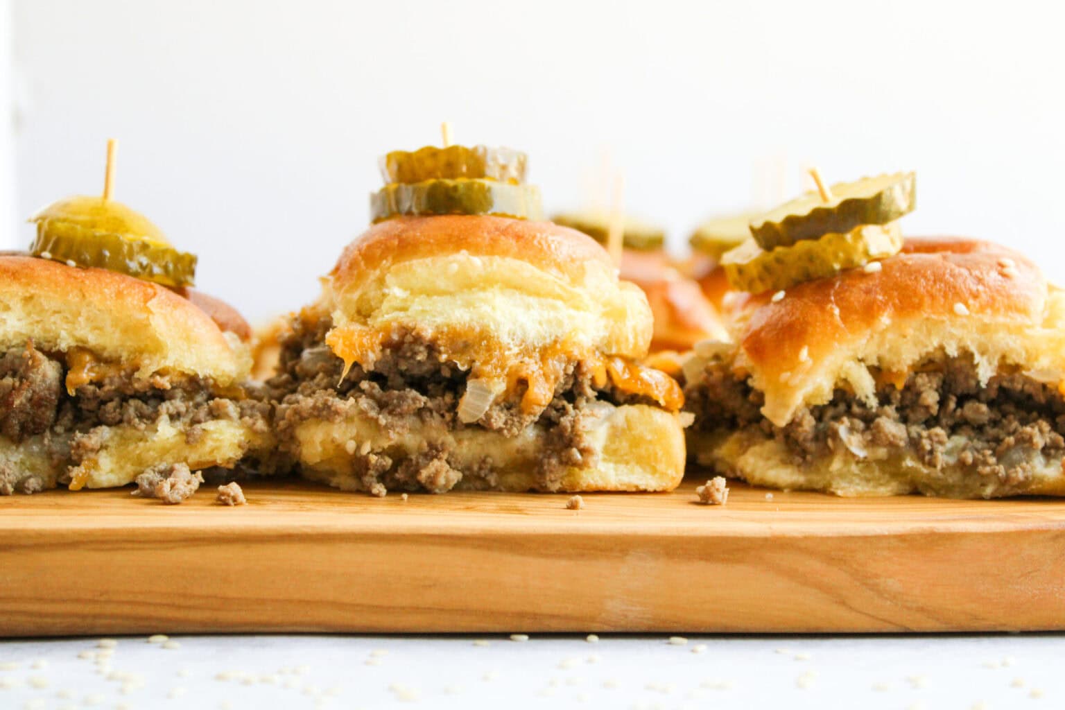 Small beef and cheese sliders topped with pickle slices on wooden board, each held together with a toothpick.