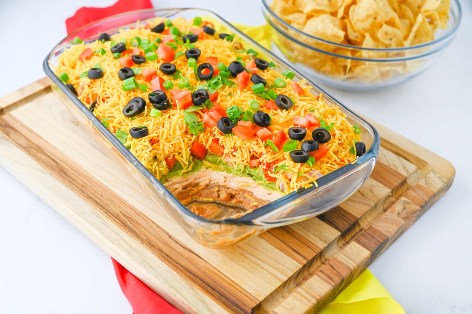 Glass dish filled with layered dip topped with shredded cheese, tomatoes, olives, and green onions, set on a wooden board next to a bowl of tortilla chips.