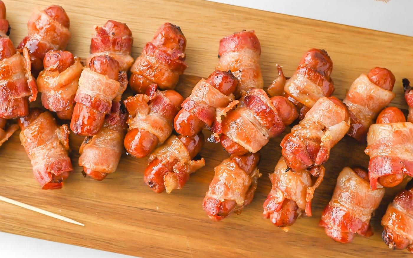 A wooden board with rows of small sausages wrapped in crispy, cooked bacon, arranged neatly for serving.