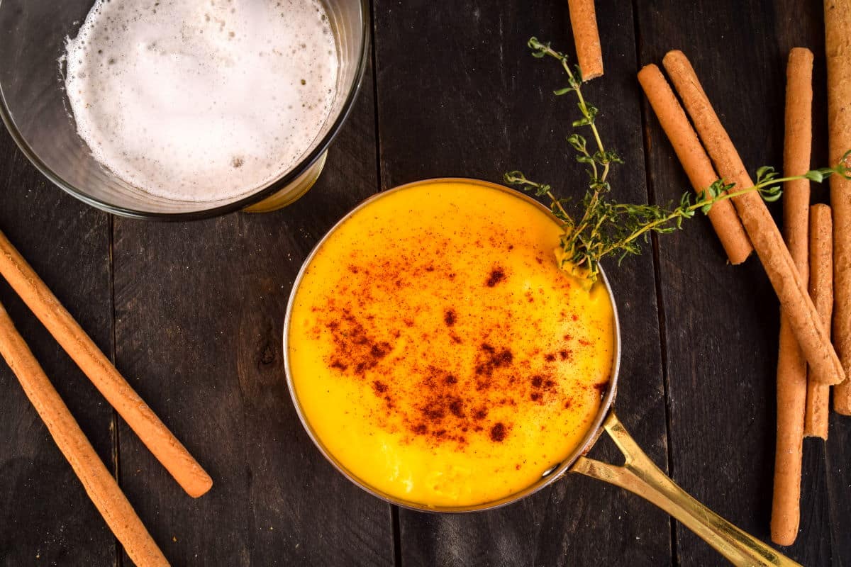 A bowl of creamy yellow soup garnished with paprika and thyme, surrounded by cinnamon sticks, next to a glass of frothy beverage on a dark wooden surface.