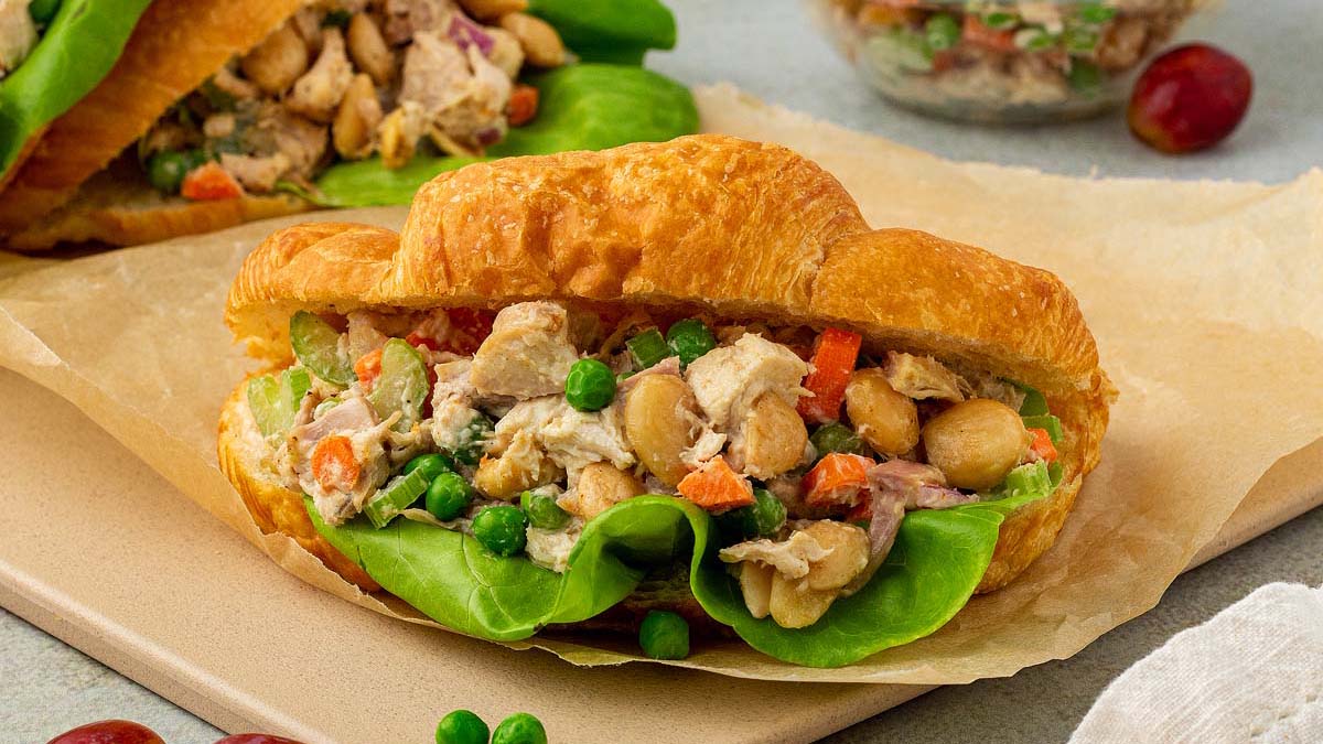 A croissant sandwich filled with lettuce, chicken salad, peas, carrots, and chickpeas, placed on parchment paper.