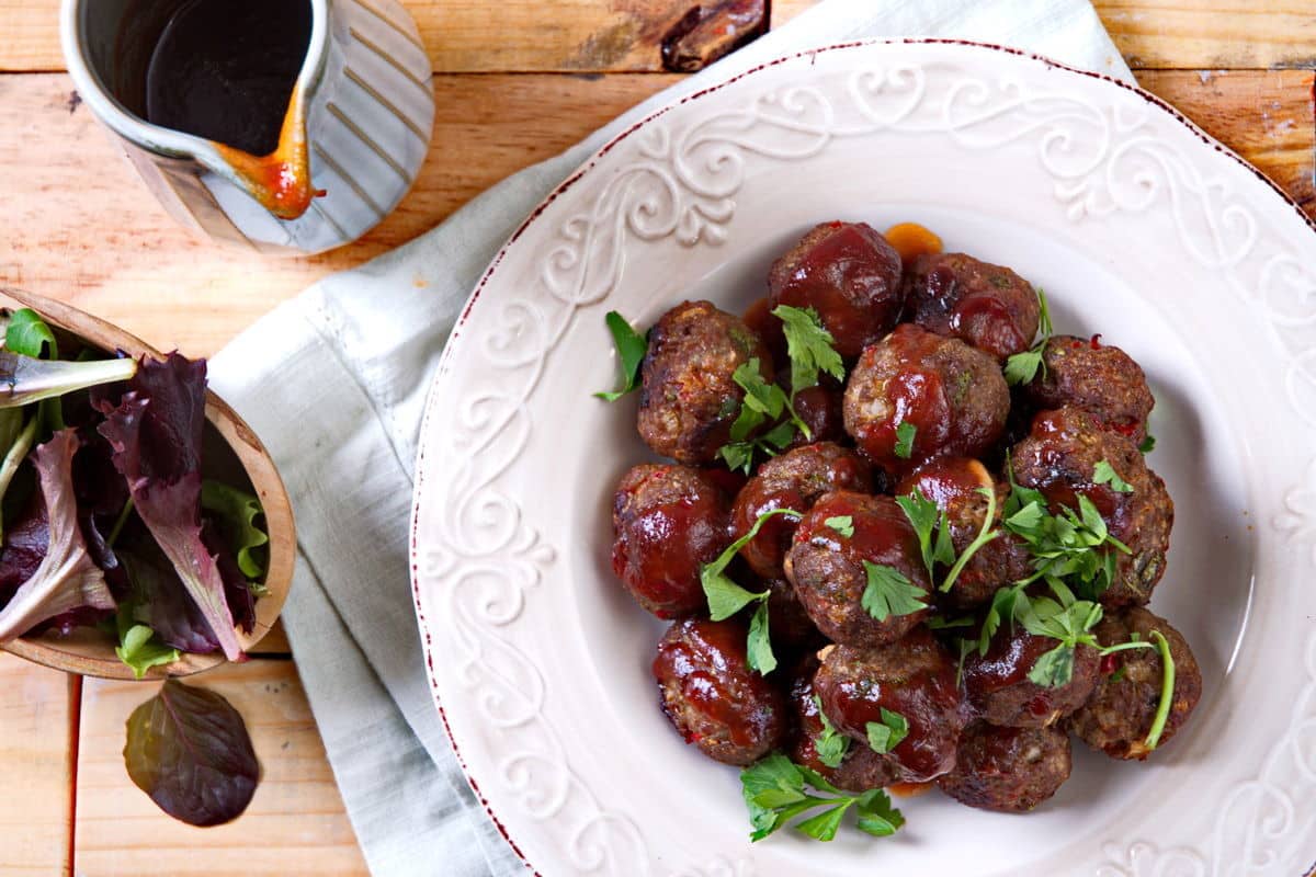A white plate filled with saucy meatballs garnished with parsley, next to a small jug of sauce and a bowl of mixed greens on a wooden table.