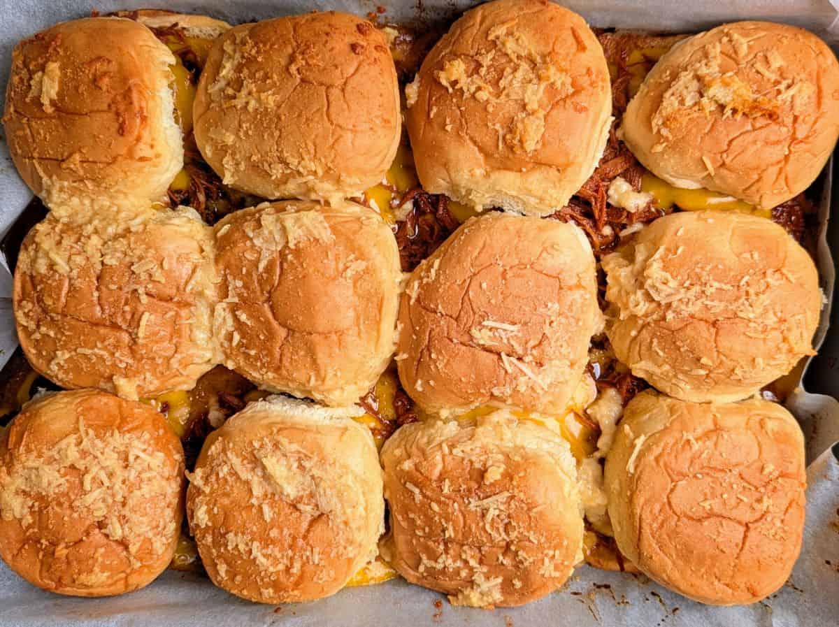 A tray of twelve baked slider sandwiches with golden brown tops, filled with cheese and shredded meat, arranged closely together.