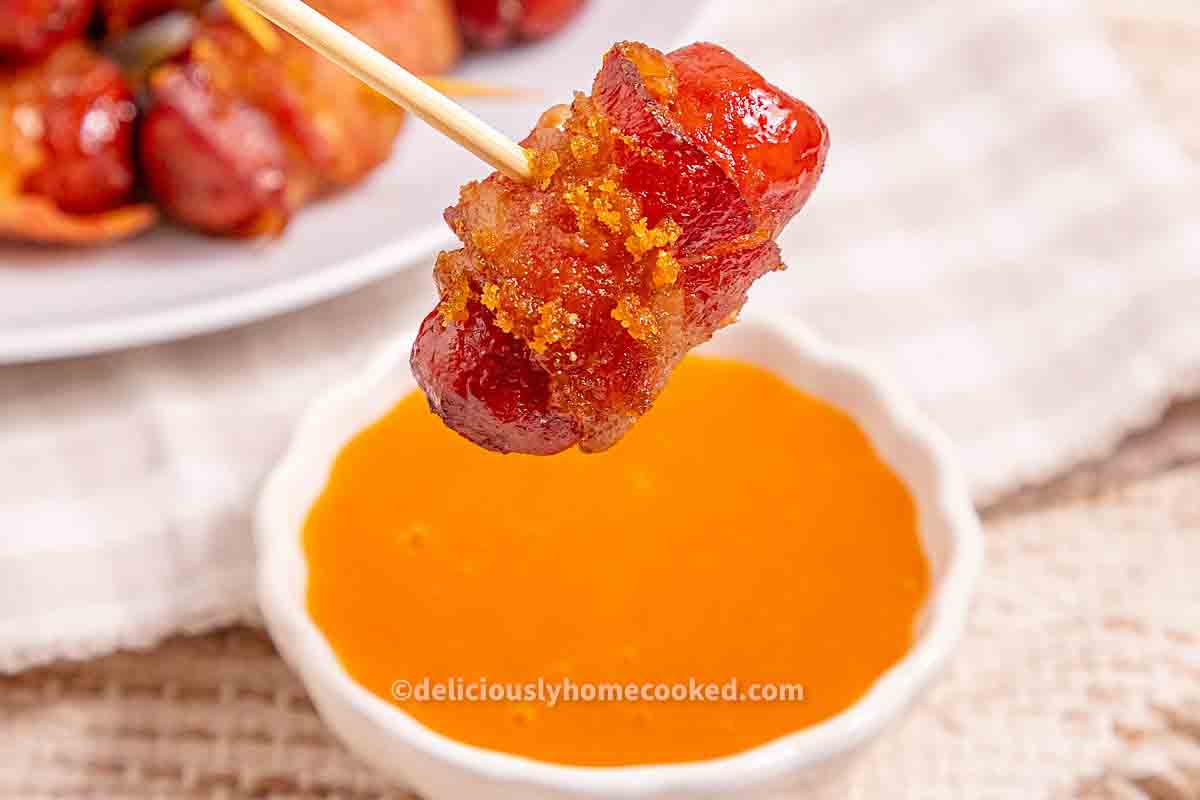 A bacon-wrapped sausage on a toothpick is held above a small bowl of orange dipping sauce.