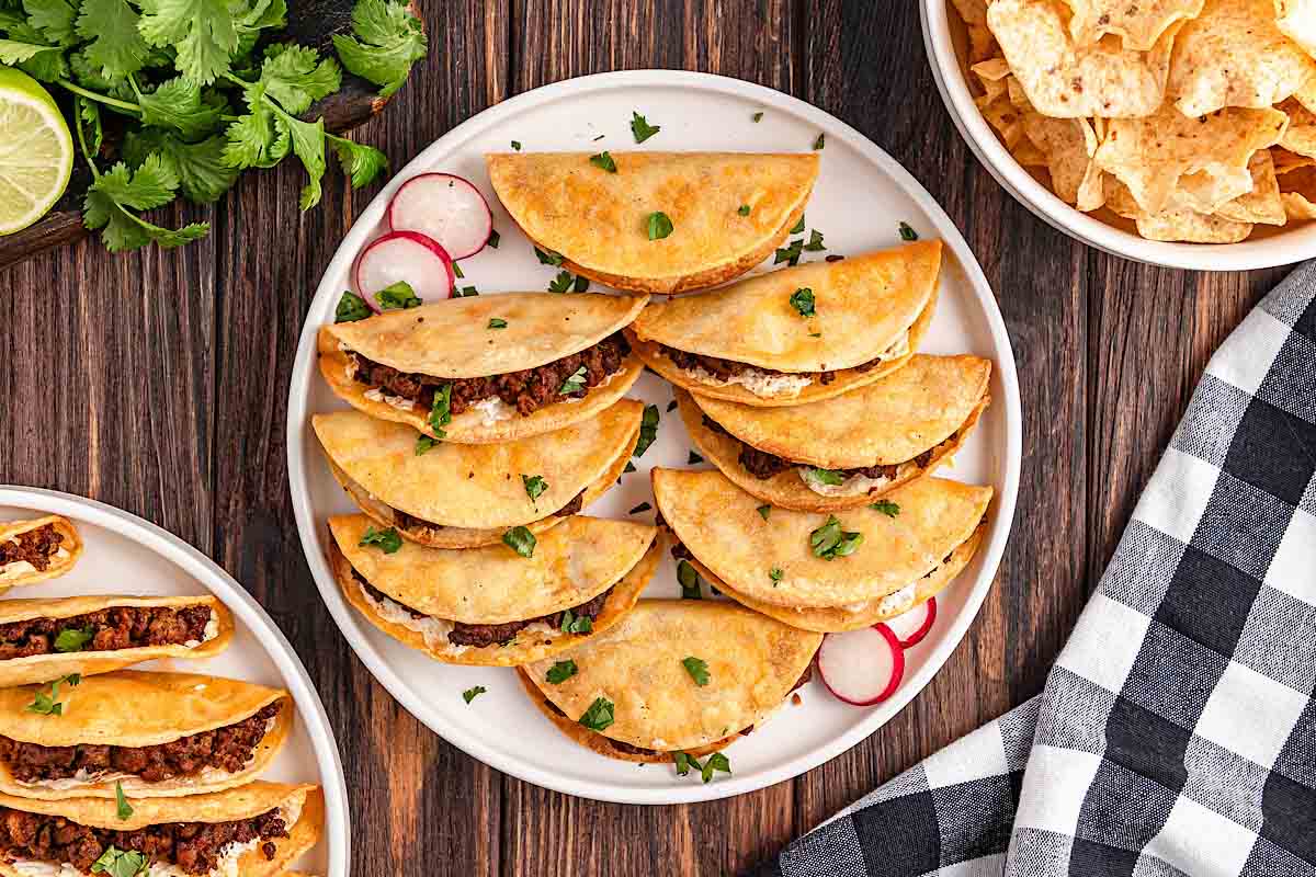 A plate of crispy tacos filled with seasoned meat, garnished with chopped cilantro and sliced radishes, next to a bowl of tortilla chips and a checkered napkin.