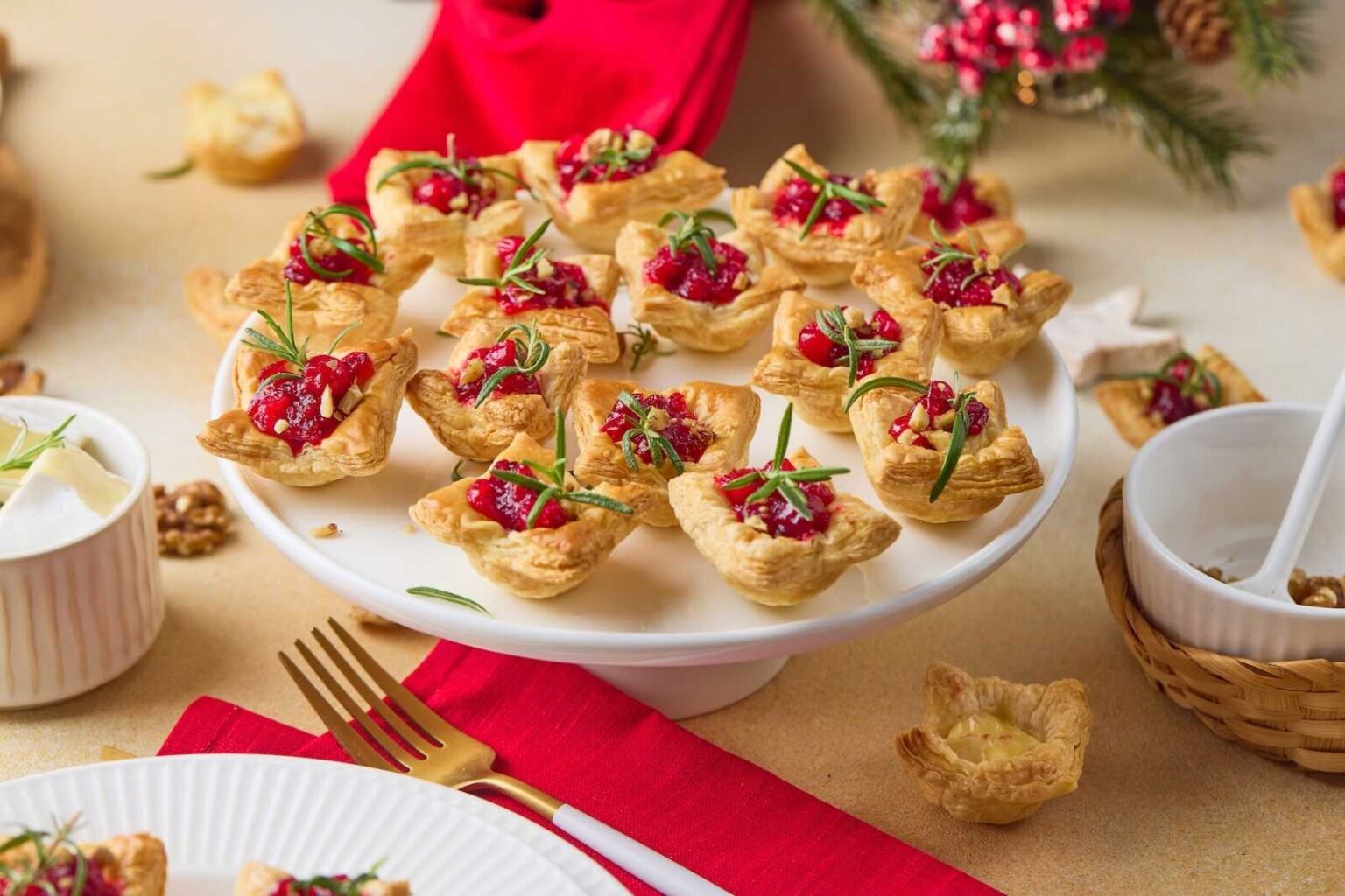 A white cake stand holds puff pastry appetizers topped with pomegranate seeds and herbs, set on a festive table with red napkins and holiday decorations.