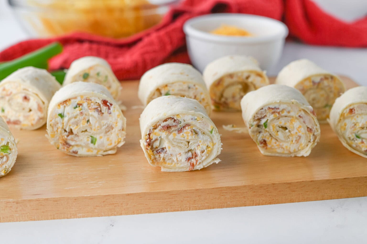 Slices of tortilla pinwheels filled with a creamy mixture, placed on a wooden board, with a bowl of dip and a red cloth in the background.