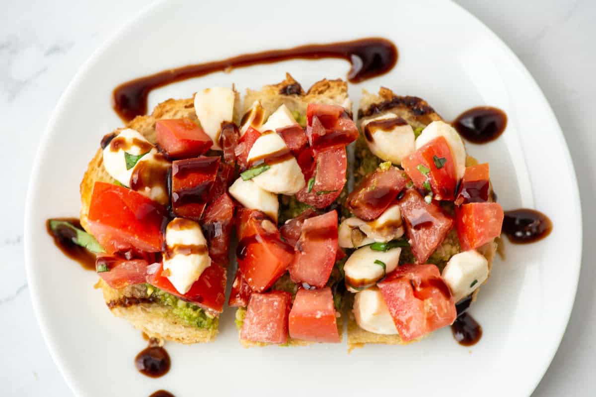 A plate with sliced bruschetta topped with chopped tomatoes, mozzarella, basil, and drizzled balsamic glaze.