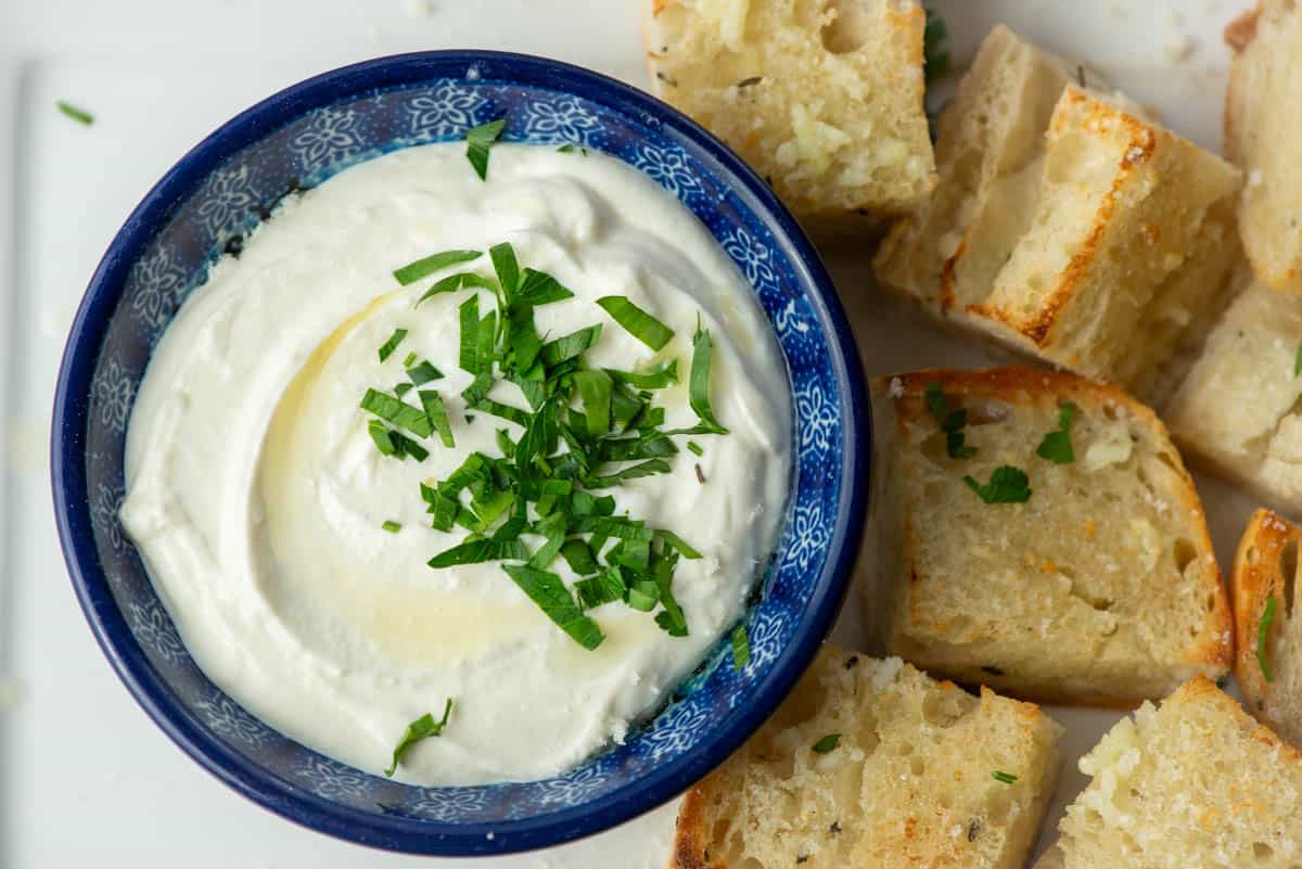 A bowl of creamy white dip garnished with chopped herbs, served with chunks of bread on the side.