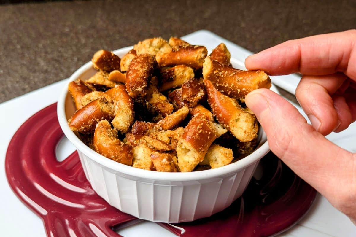 A hand reaches for a piece of seasoned pretzel bites in a white bowl, set on a maroon silicone mat.