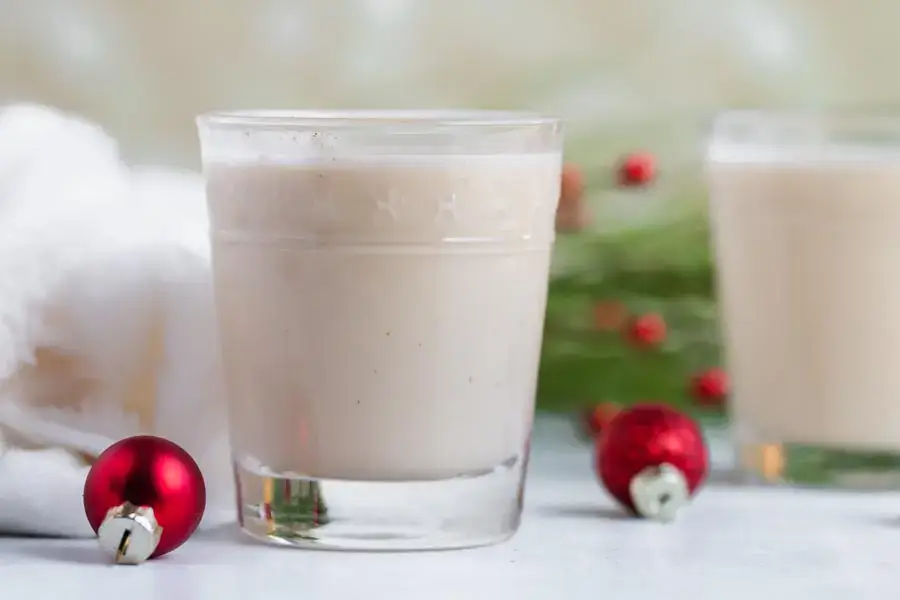 A glass of creamy eggnog sits on a light surface, with red Christmas ornaments and greenery in the background.