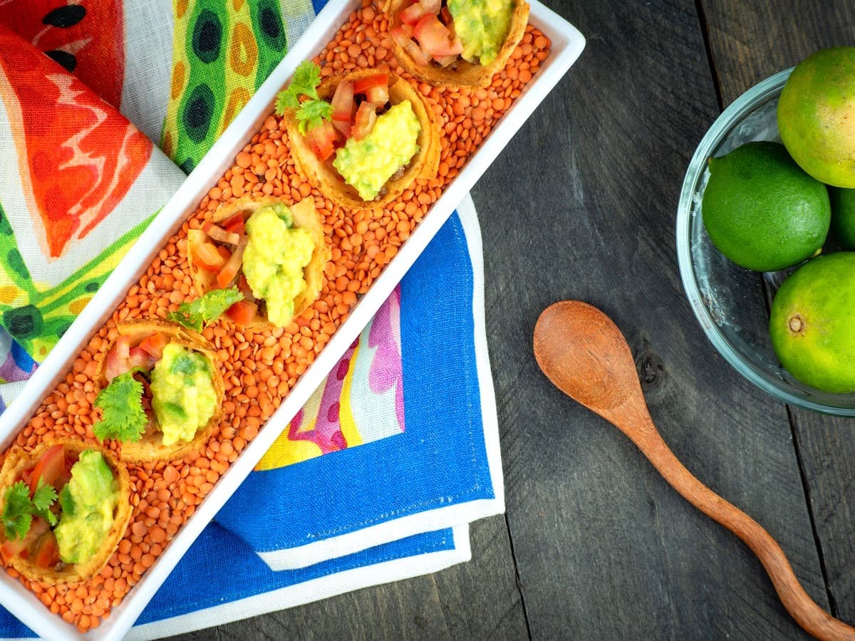 A rectangular white dish with five mini taco cups filled with guacamole and salsa on lentils, next to a bowl of limes, wooden spoon, and colorful napkins on a wooden table.