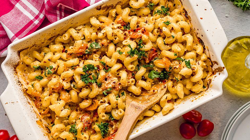 Baked pasta casserole with curly noodles, cheese, and herbs in a white dish, mixed with a wooden spoon. Red napkin and cherry tomatoes are nearby.