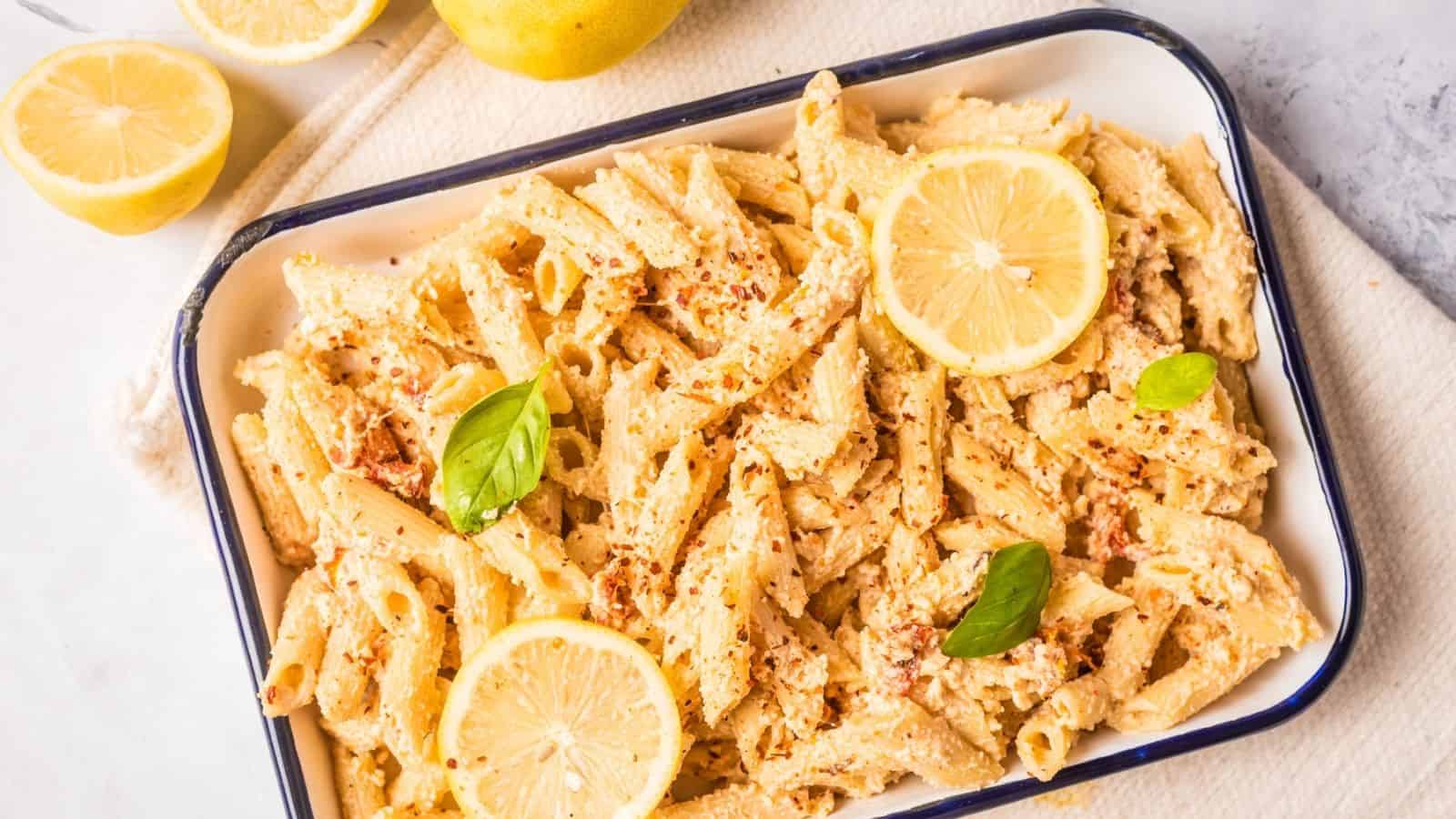 Baked penne pasta in a white dish, garnished with lemon slices, basil leaves, and sprinkled with seasoning. Halved lemons are placed beside the dish.