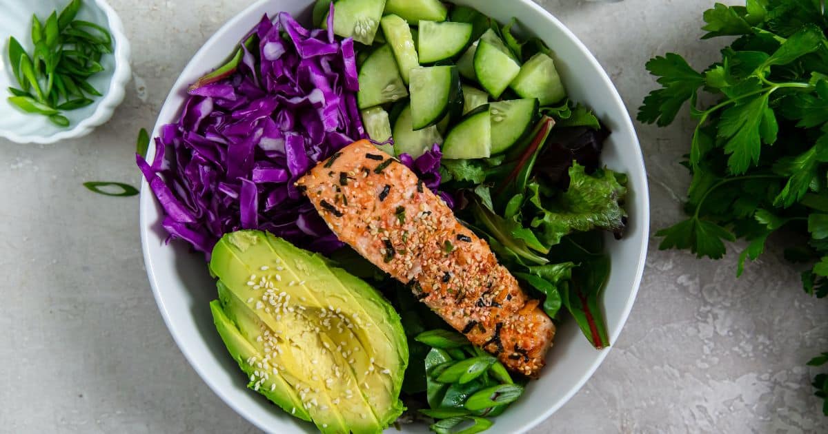 A bowl containing sliced avocado, purple cabbage, cucumber, mixed greens, green onions, and a seasoned salmon fillet.