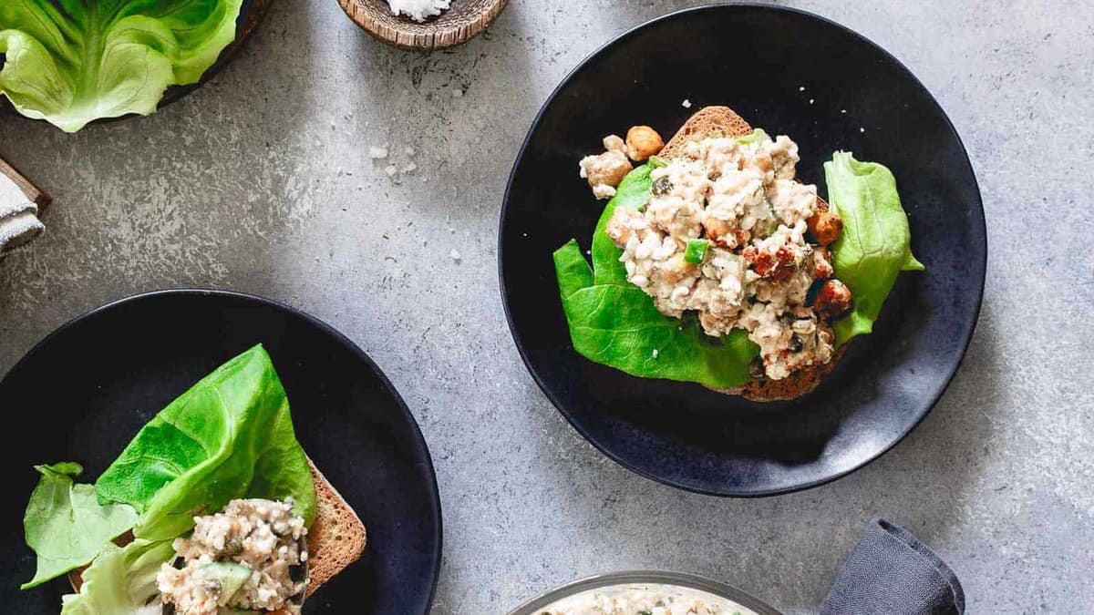 Two black plates with open-faced sandwiches featuring leafy lettuce and a creamy salad mixture on brown bread, set on a gray surface.
