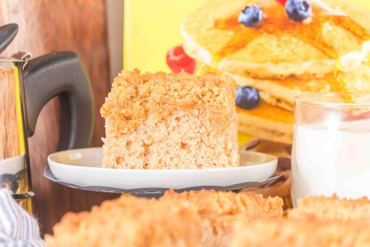 A slice of crumb cake on a plate, with a stack of pancakes topped with blueberries and a glass of milk in the background.