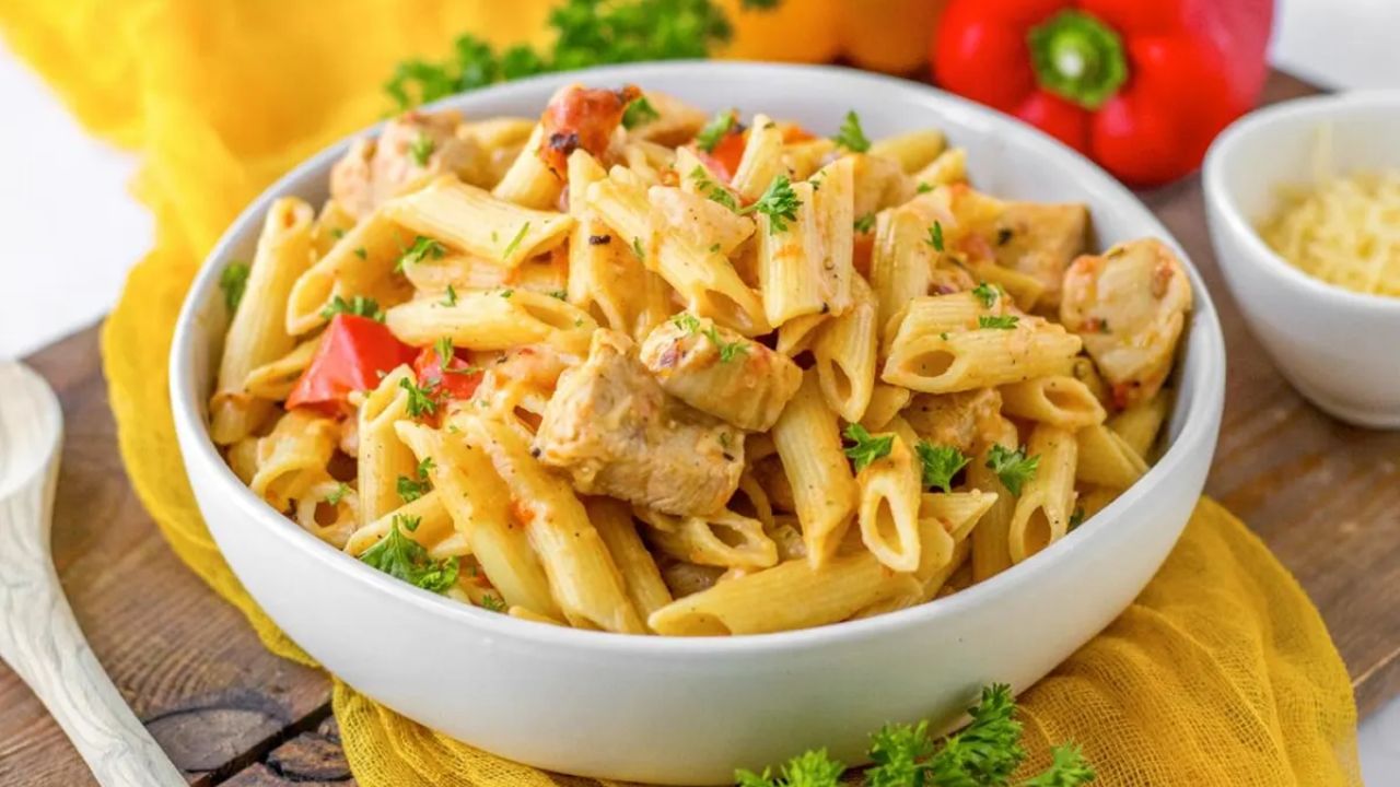 A white bowl filled with penne pasta, diced chicken, red bell peppers, and parsley is set on a wooden surface with a small bowl of shredded cheese and vegetables nearby.