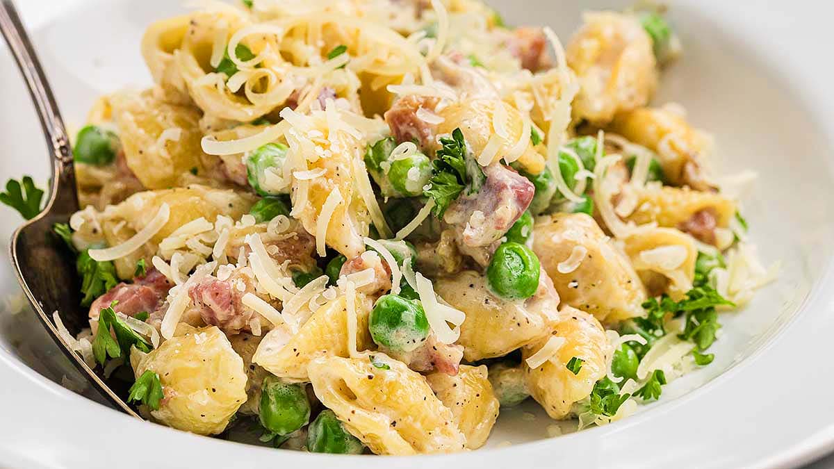 A plate of creamy pasta with peas, diced ham, shredded cheese, and parsley, served with a spoon.