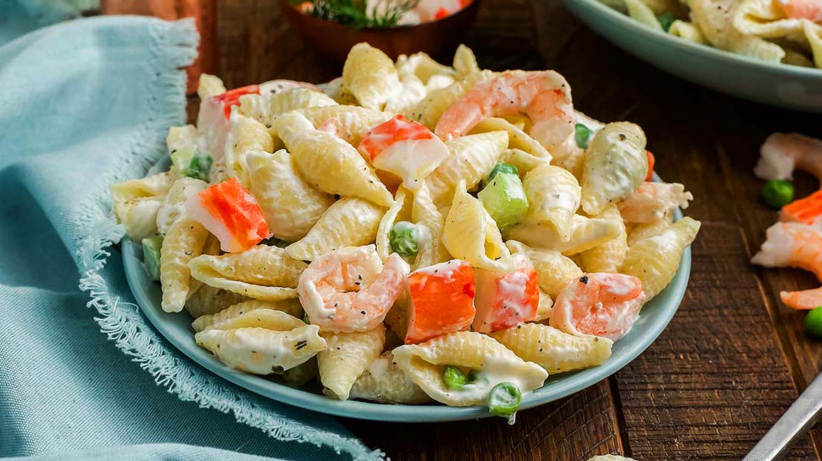 A plate of shell pasta salad with shrimp, imitation crab, green peas, and creamy dressing, served on a wooden table with a blue napkin nearby.